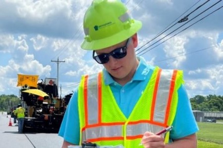 An image of a civil engineer trainee with yellow hard hat and yellow safety vest and black sunglasses at a construction site with a clipboard in one hand and a pen in the other hand with a large piece of equipment and other workers in the background